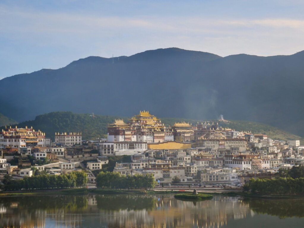Panoramic view of Songzanlin Monastery (Ganden Sumtseling Monastery), the largest Tibetan Buddhist monastery in Yunnan Province, nestled in Shangri-La (Dêqên), China. The grand golden-roofed temple complex with its distinctive white buildings spreads across the hillside against dramatic blue mountain ranges, with reflections visible in the foreground lake at golden hour.